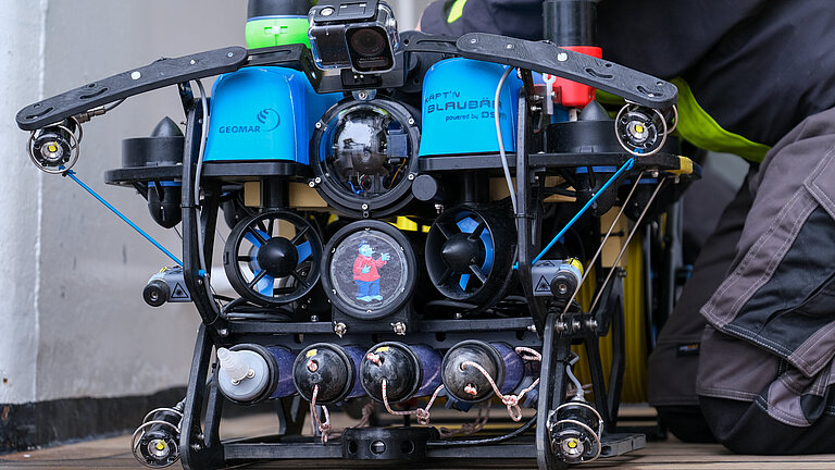 A device with a camera eye, two blue lids and a sticker in the centre with the picture book character Captain Bluebear on it stands on the deck of a ship