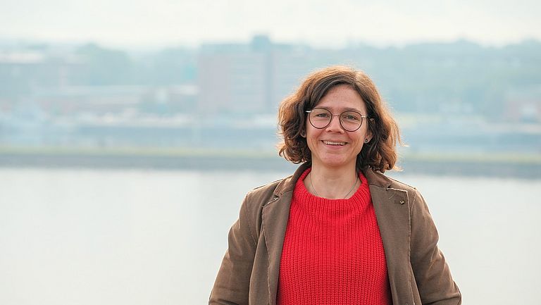 Eine junge Frau steht vor einer Wasserfläche, im Hintergrund Hafenanlagen
