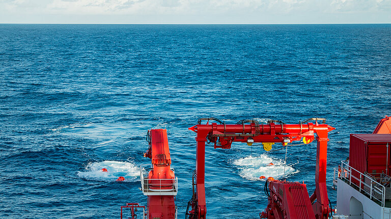 Blaues Meer, unten rechts im Bild sind der rote Kran eines Forschungsschiffes zu sehen, hinter dem Schiff werden zwei rote Bojen geschleppt