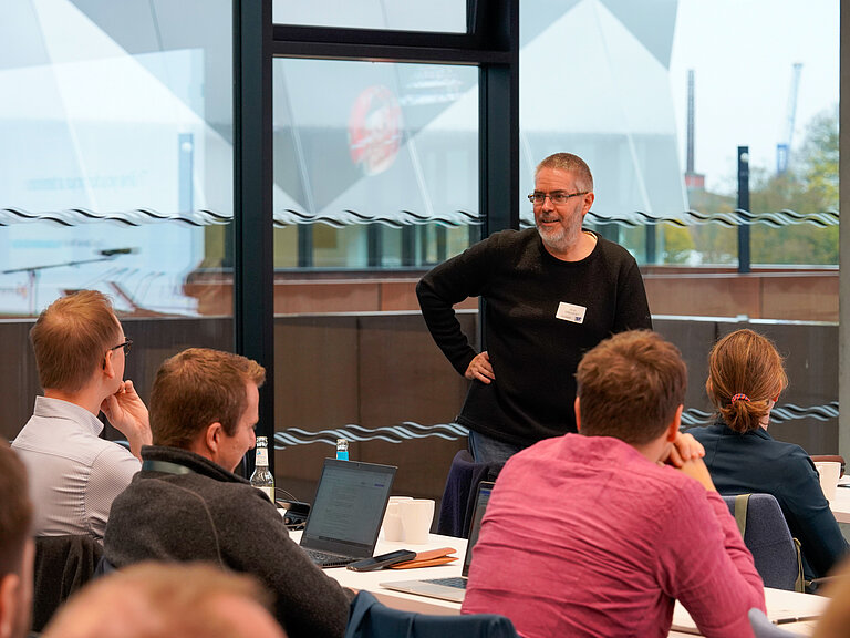 A man with glasses and short grey hair speaks to a group of listeners at tables in a conference room