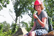 Eine junge Frau mit einem roten Schutzhelm sitzt auf einem großen Stein