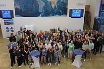 Eine Gruppe Menschen steht in einem Foyer. Einige von ihnen zeigen auf einen Bildschirm mit der Aufschrift "Stronger together".